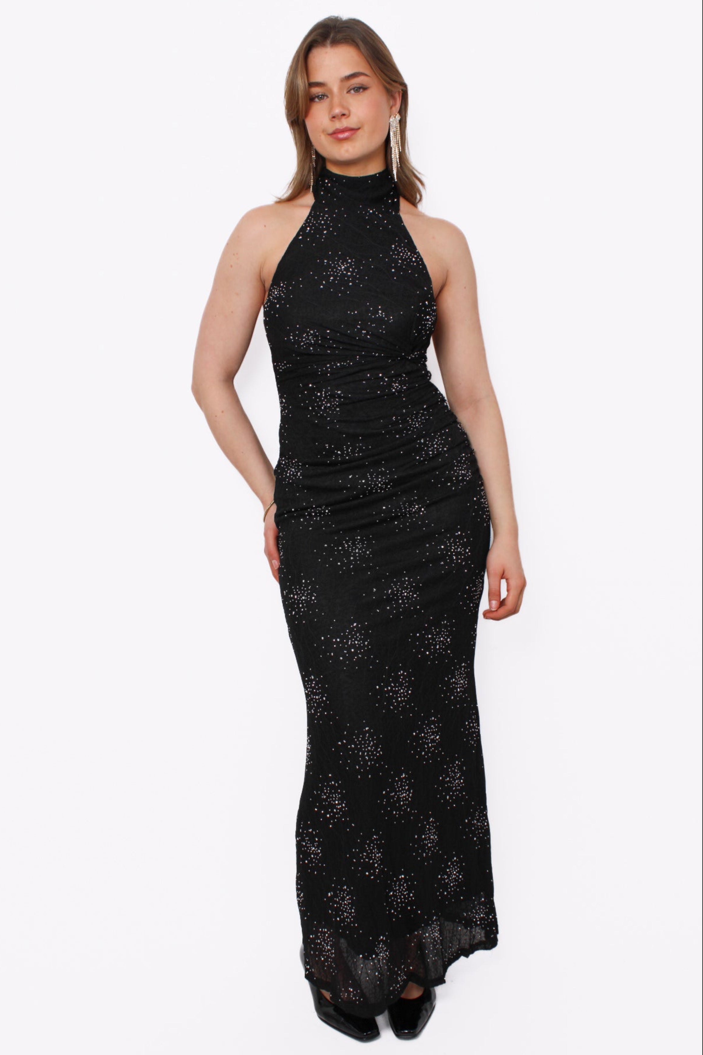 Deze zwarte maxi-jurk straalt pure elegance uit dankzij de subtiel glinsterende details die het hele silhouet laten sprankelen. De hoge halternek creëert een verfijnde, vrouwelijke look, terwijl de zachte plooien in de taille de pasvorm mooi laten aansluiten. De echte verrassing bevindt zich aan de achterkant: een open rug die de jurk een sensuele, maar chique twist geeft. De luchtige stof valt soepel langs het lichaam en beweegt elegant met je mee. Draag deze jurk met opvallende oorbellen en klassieke hakk